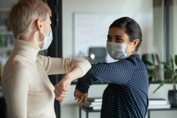 Smiling diverse female colleagues wearing protective face masks greeting bumping elbows at workplace, woman coworkers in facial covers protect from COVID-19 coronavirus in office, healthcare concept